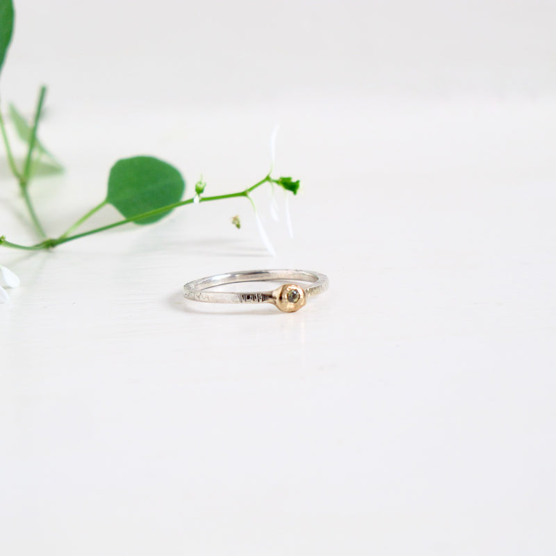 Image shows a delicate ring in silver and gold. The fine band of 1.50 mm is textured with horizontal lines all the way round . Sitting on the top of the band is a nugget-like 14 Kt gold bauble with a green sapphire flush set into it the centre of it. 