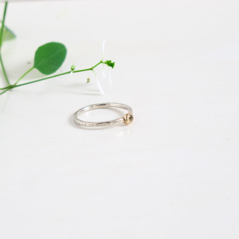 Image shows a delicate ring in silver and gold. The fine band of 1.50 mm is textured with horizontal lines all the way round . Sitting on the top of the band is a nugget-like 14 Kt gold bauble with a green sapphire flush set into it the centre of it. 