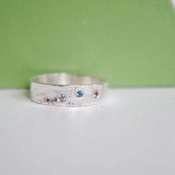 A  6 mm wide silver band with an organic looking hand textured surface.  Three faceted sapphires of different colours are set in a scattered arrangement that is pleasing to the eye. 