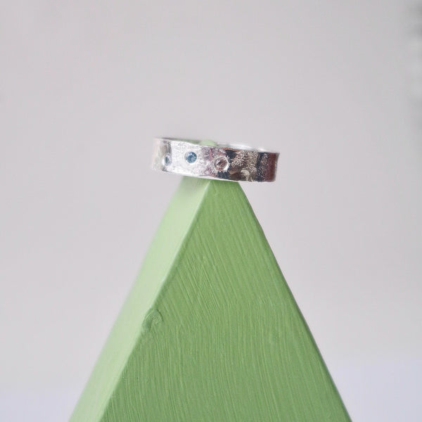 A  6 mm wide silver band with an organic looking  surface that is custom textured by hand with unique stamps and a rolling mill.  Three faceted sapphires of different colours are set in a scattered arrangement that is pleasing to the eye. 