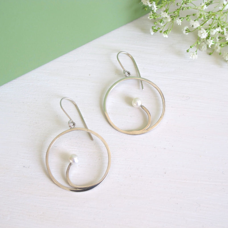 A  genuine round freshwater 5 mm  white pearl sits within a silver crescent segment that is inside a handcrafted silver hoop earring.