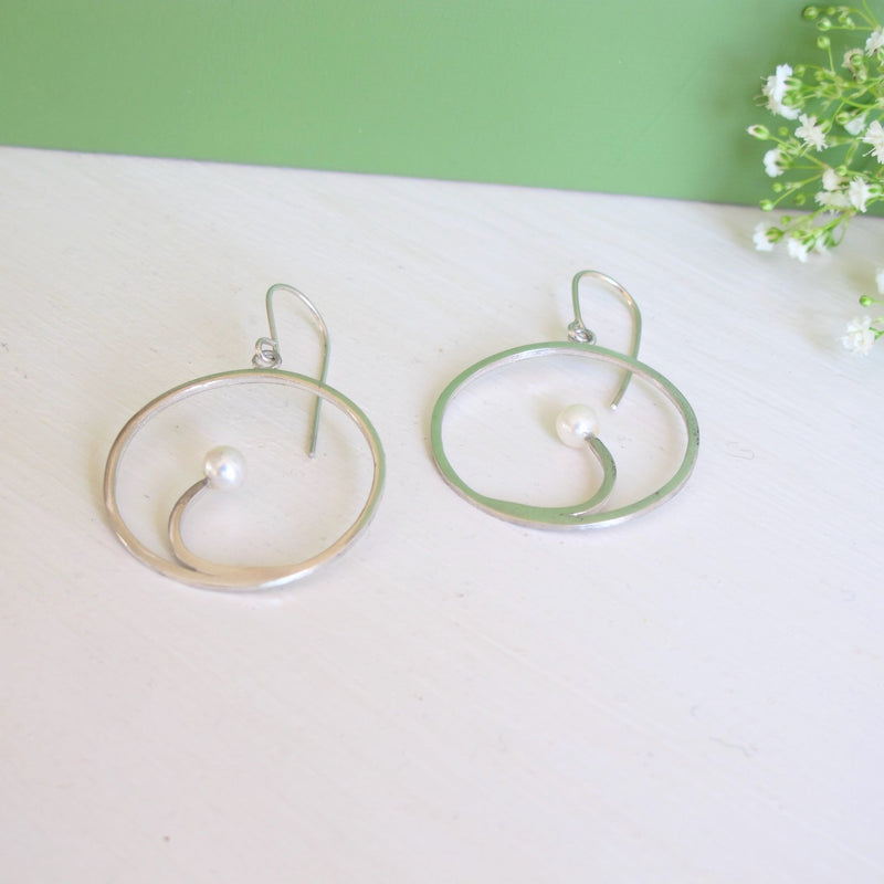 A  genuine round freshwater 5 mm  white pearl sits within a silver crescent segment that is inside a handcrafted silver hoop earring.