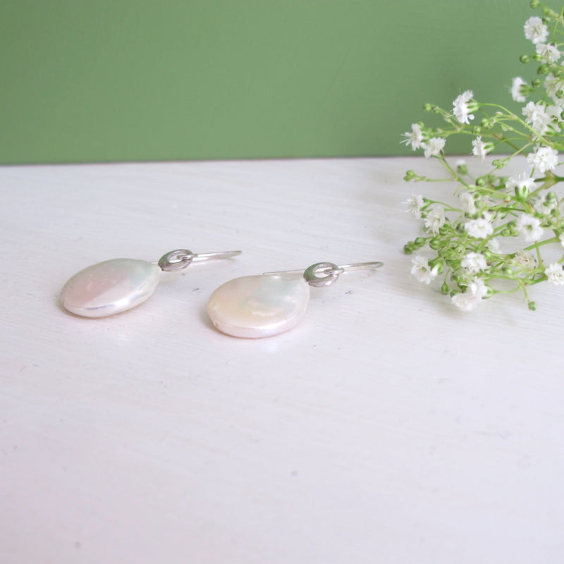 An image of a large white freshwater teardrop shaped coin pearl. At the top of the pearl sits a unique small semi oval silver finding which the ear hook  threads through and the pearl articulates from. 