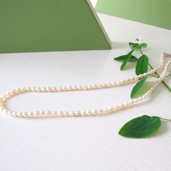 A closeup of a hand strung genuine pearl necklace  composed of small  
 white luminous oval freshwater pearls.