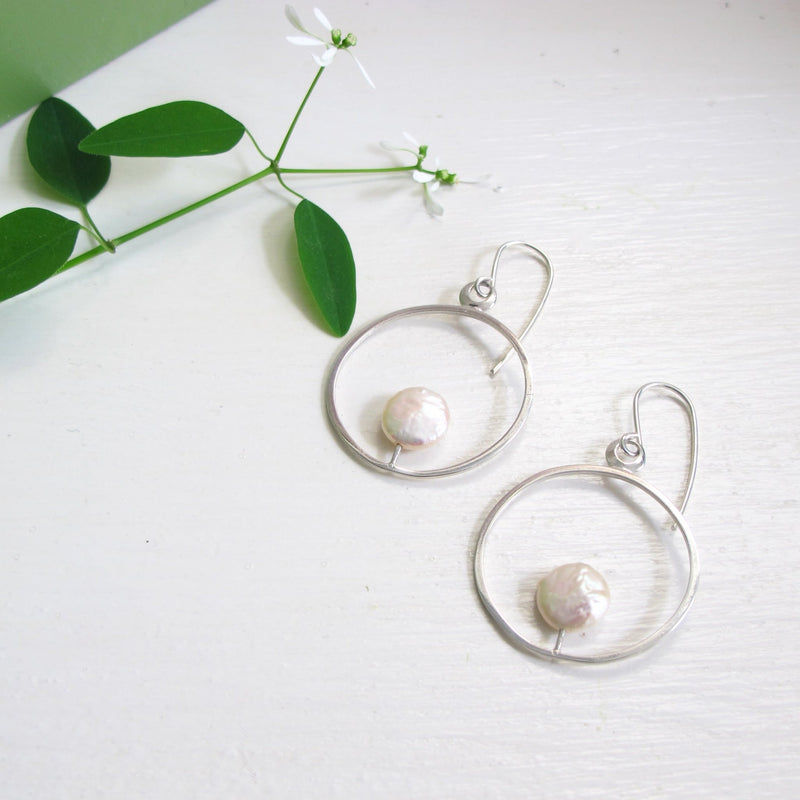 A pair of mid size round silver reiki imbued hoop earrings with a consciously sourced genuine round white coin pearl that sits at the tip of a fine silver post that is attached to the inside base of the hoop.