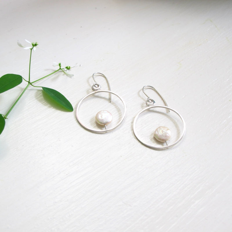 A pair of mid size round silver reiki imbued hoop earrings with a consciously sourced genuine round white coin pearl that sits at the tip of a fine silver post that is attached to the inside base of the hoop.