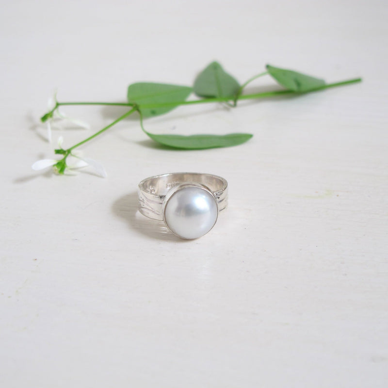 A 11.65 mm freshwater white pearl holds the viewer's eye. IT is set into a sterling silver bezel and sits on a 5.60mm silver band which has hand engraved  flowing lines that wrap around the band adding a unique detail to this ring. A one-of  a kind beauty. 