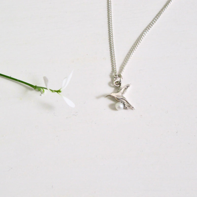 A 10 mm Hummingbird depicted in flight hangs from a silver chain.  A tiny pearl sits at the bottom of the bird's breast.