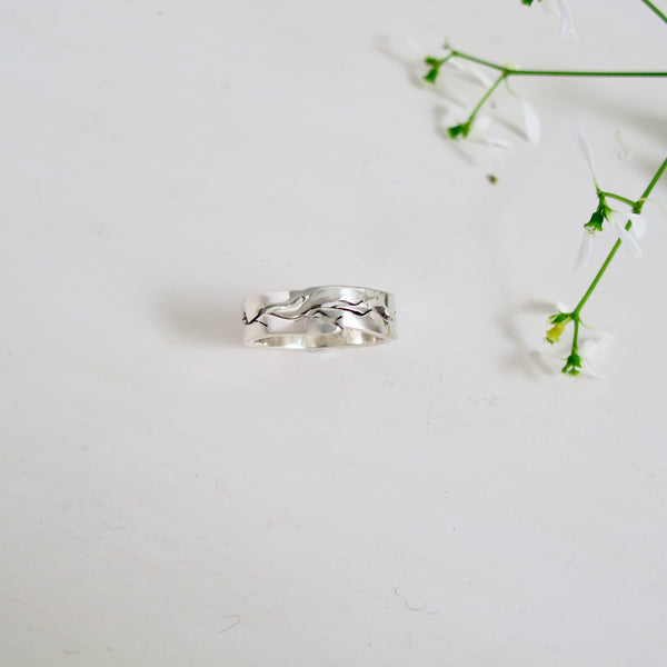 A 6 mm wide sterling silver band  with undulating edges. The centre of the ring has cut out thin branch-like design that wrap around the band. 