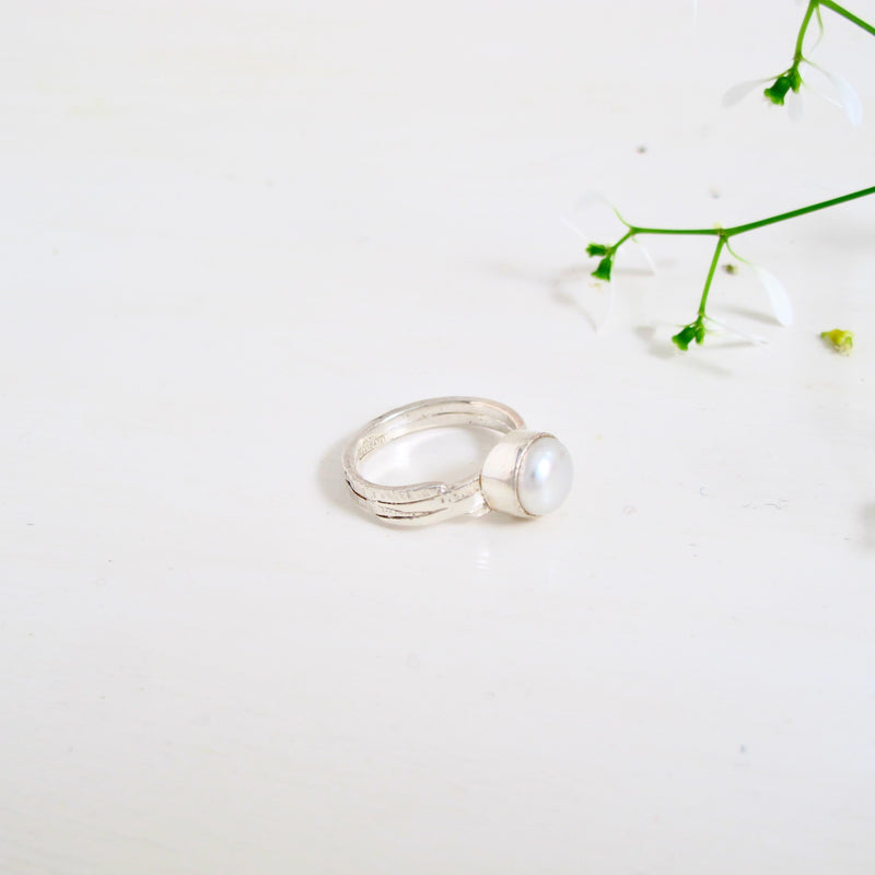 A sterling silver ring with a large 10 mm white freshwater pearl that set in a sterling silver bezel sits on a 6 mm wide silver band. The band of the ring is hand engraved with organic lines that work around the band.  Between the lines are scattered with small textural details like tiny circles that resemble tiny bubbles. 