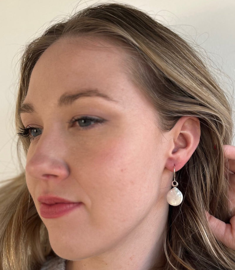 From a woman's earlobe drops a large white round coin pearl with a unique finding for the earhook to attach to the earrings