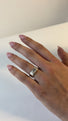 A woman places a narrow  wedding band onto her finger next to a wider cushion cut diamond engagement ring. 