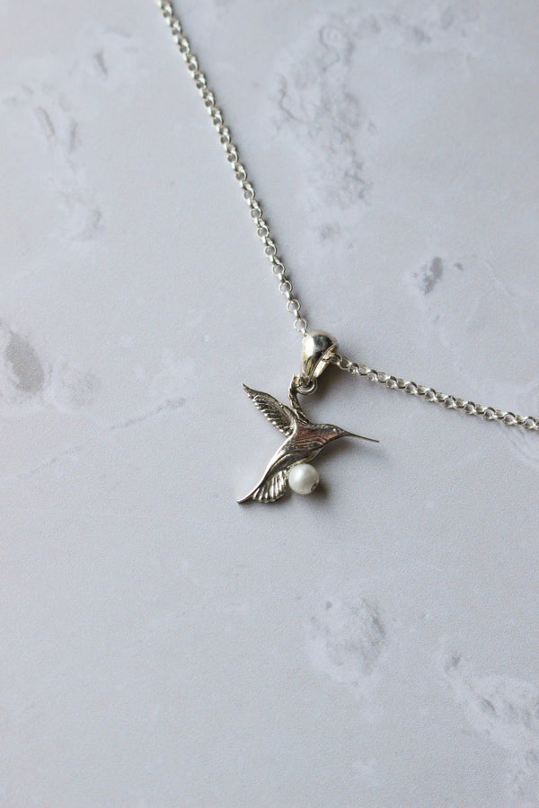 A 10 mm Hummingbird depicted in flight hangs from a silver chain.  A tiny pearl sits at the bottom of the bird's breast.