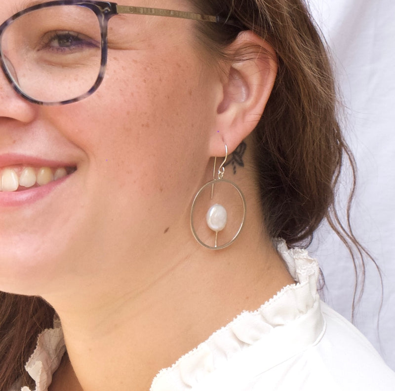 Close up of a young woman’s neck and ear, showing a delicate Reiki imbued handcrafted hoop earring of consciously sourced sterling silver containing a large flat white coin pearl 1/3 the size of the hoop dangling midway between ear and shoulder.