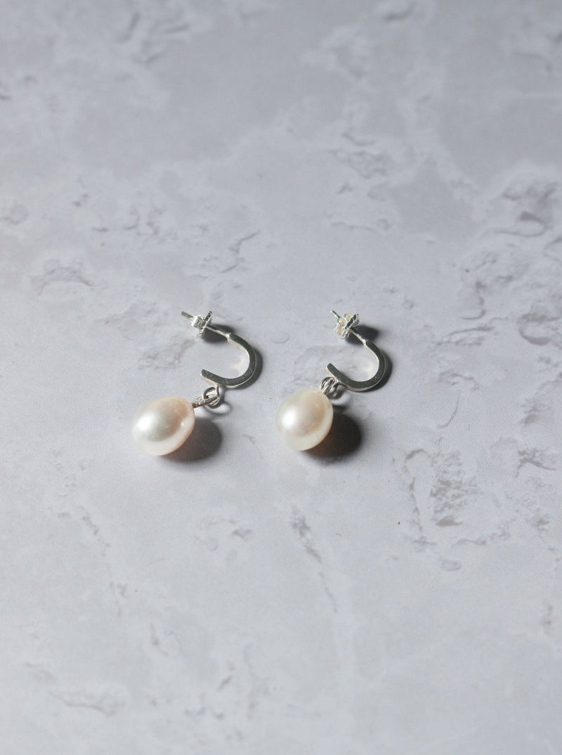A pair of silver earrings with a white oval pearl dropping from a curved crescent with an ear post at the end of the crescent