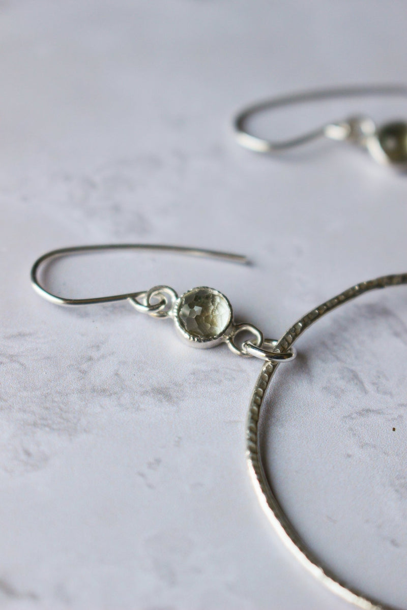 An oversized Reiki Imbued textured silver hoop with a 6 mm rose cut green amethyst that links the hoop and ear hook together.