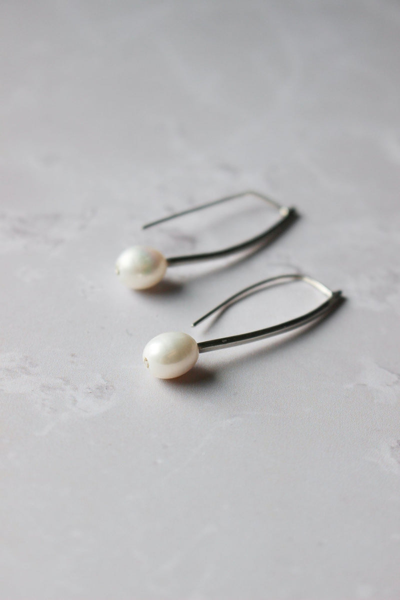 An oval consciously sourced white pearl sits at the end of a curved silver bar that dangling from the ear.