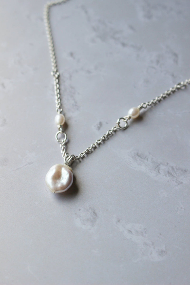 A white Baroque pearl hangs from a silver chain. The chain is embellished with two small oval white pearls near the centre.