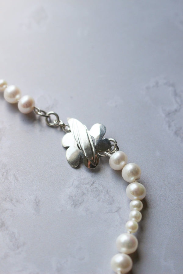 A closeup of various sizes of white pearls hand strung on silk to create necklace with a 6 petal original silver clasp.