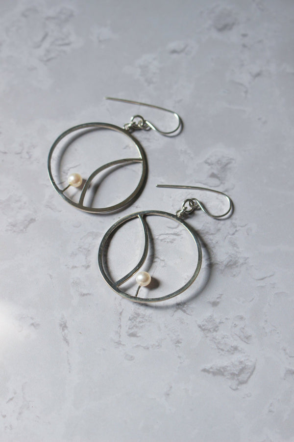 A freshwater white pearl sits within a silver crescent segment that is inside a handcrafted hoop earring imbued with Reiki.